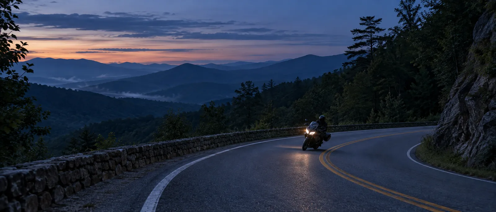 A motorcycle with its headlight on leans gently through a curve on a mountain road at blue hour.